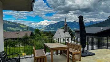 Große Terrasse mit Holztisch und Bänken, bietet einen weiten Blick auf die Berge und eine Kirche.