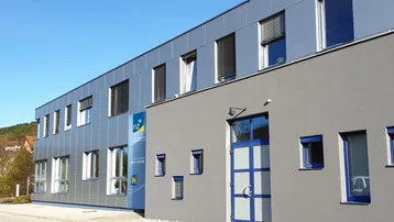 Moderne Bürogebäude-Fassade mit grauen Paneelen und blauen Fensterrahmen unter klarem Himmel.