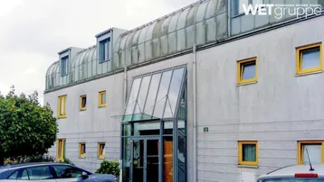 Ansicht der modernen Gebäudefassade mit gelben Fensterrahmen und Glasvordach am Eingangsbereich.