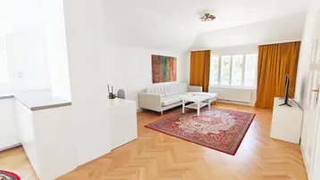 Helles Wohnzimmer mit Fischgrätparkett, beiger Couch und farbigen Vorhängen, offen zum Küchenbereich.