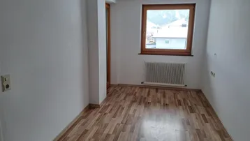 Helles Zimmer mit Laminatboden, Heizkörper und Fenster mit Blick auf die Berge.