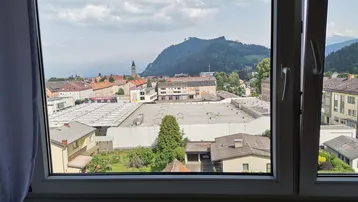Ausblick aus dem Fenster auf die Stadt mit Gebäuden und grünen Bergen im Hintergrund.