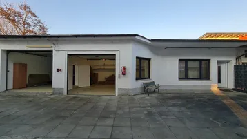 Außenansicht eines Gebäudes mit zwei Garagentoren und einem gepflasterten Vorplatz bei Tageslicht.