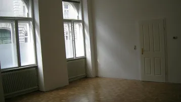 Heller Raum mit Parkettboden, hohen Fenstern und weißer Tür.