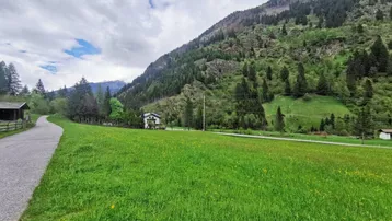 Weitläufige grüne Wiese mit einem Schotterweg und Blick auf bewaldete Berge unter bewölktem Himmel.