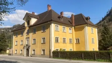 Historisches Gebäude mit gelber Fassade und braunem Dach, umgeben von grüner Landschaft und Bergen.