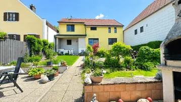 Großer Garten mit Rasenfläche, Pflanzen und einem gemauerten Grillbereich hinter dem gelben Haus.