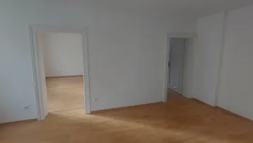 Leerer Raum mit hellem Holzfußboden und weißen Wänden, der Zugänge zu weiteren Zimmern bietet.