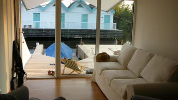 Helles Wohnzimmer mit großen Fenstern, Holzboden und Blick auf die Holzterrasse und die Wasserhäuser.