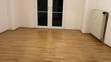 Leeres Zimmer mit hellem Holzfußboden und Zugang zu einem Balkon oder einer Terrasse.