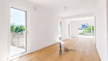 Lichtdurchfluteter Raum mit Holzboden, großen Fenstern und Zugang zu einem Balkon.