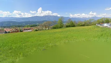 Weitläufige grüne Wiese mit Blick auf die umliegende Berglandschaft und eine Siedlung.