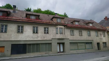 Altes Gebäude mit mehreren Fenstern und Ladenfronten an einer Straße unter bewölktem Himmel.