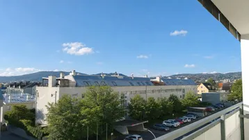 Blick vom Balkon auf die umliegende Bebauung und die grünen Hügel im Hintergrund unter blauem Himmel.