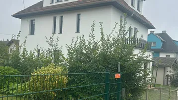 Weißes Wohnhaus mit Satteldach, umgeben von einem grünen Zaun und gepflegten Büschen.