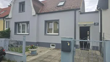 Graues Einfamilienhaus mit Mansarddach, weiß gestrichenen Fenstern und einem kleinen Vorgarten.