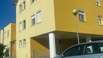 Gelbes Mehrfamilienhaus mit schräger Dachlinie und geparktem Auto davor.
