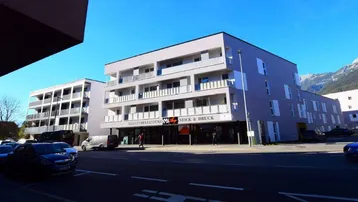 Modernes Geschäfts- und Wohngebäude mit großen Fenstern und Balkonen an einer belebten Straße mit Bergblick.