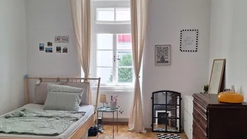 Helles Zimmer mit Holzboden, einem Bett, Sofa und einem Fenster mit Vorhängen.