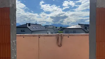Blick aus einem Fensterrahmen auf moderne Neubauten und die umliegende Berglandschaft unter blauem Himmel.