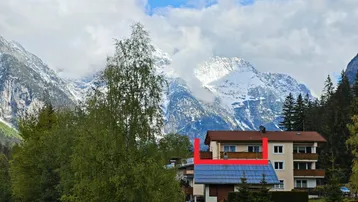 Mehrfamilienhaus in alpiner Umgebung mit schneebedeckten Bergen im Hintergrund und grüner Vegetation.