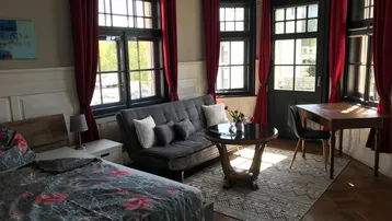 Geräumiges Wohnzimmer mit Parkettboden, bequemer Couch, Schreibtisch und großen Fenstern für viel Tageslicht.