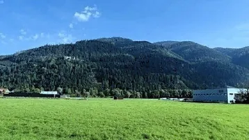 Weitläufiges grünes Feld mit bewaldeten Bergen im Hintergrund unter blauem Himmel.