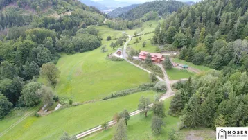 Luftaufnahme eines Anwesens mit Haupthaus und Nebengebäude, eingebettet in grüne Hügel und Wälder.