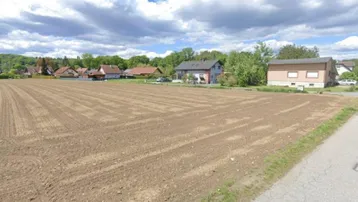 Weitläufiges Ackerland mit Wohnhäusern im Hintergrund unter blauem Himmel und Wolken.