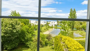 Weitblick aus dem Fenster auf einen Teich, grüne Bäume und moderne Häuser.