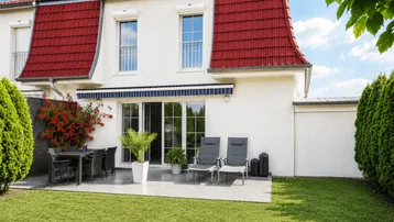 Hinteransicht eines modernen Hauses mit großer möblierter Terrasse, gepflegtem Garten und rotem Mansarddach.