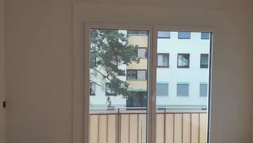 Leeres Zimmer mit hellem Holzfußboden und Zugang zu einem französischen Balkon.
