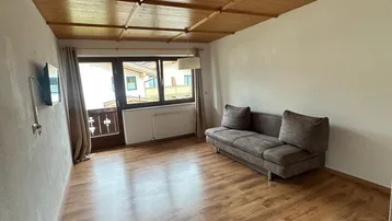 Gemütliches Wohnzimmer mit braunem Sofa, Holzbalkendecke und Zugang zu einem kleinen Balkon mit Bergblick.