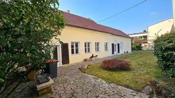 Hausfassade mit Steinterrasse und Rasenfläche im Gartenbereich unter blauem Himmel.