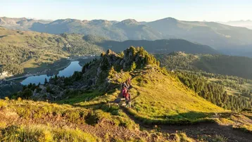 Atemberaubender Panoramablick auf eine weite Berglandschaft mit einem glitzernden See und Wanderwegen unter strahlendem Sonnenschein.