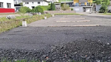 Ein Schotterparkplatz im Bau mit Blick auf bewaldete Hügel und umliegende Häuser in ländlicher Umgebung.