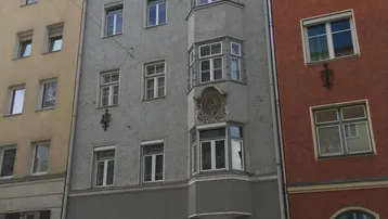 Historisches Stadthaus mit grauer Fassade, Erker und Satteldach in einer belebten Straße.