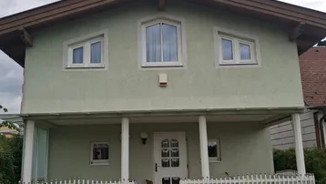 Vorderansicht eines charmanten Hauses mit hellgrüner Fassade, braunem Dach und einer überdachten Veranda.