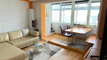 Helles Wohn- und Esszimmer mit beigem Sofa, Holzboden und einem erhöhten Essbereich an den Fenstern.