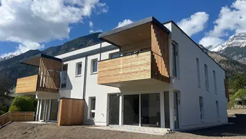 Modernes Mehrfamilienhaus mit Holzverkleidung an den Balkonen und Bergblick unter blauem Himmel.