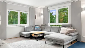 Helles Wohnzimmer mit großem Ecksofa, Teppich und Blick ins Grüne durch die Fenster.