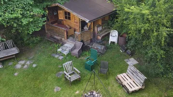 Rustikale Holzhütte in einem grünen Garten mit Feuerstelle und verschiedenen Sitzgelegenheiten.