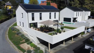 Modernes Einfamilienhaus mit Photovoltaikanlage, großer Terrasse und Carport, umgeben von Grün.