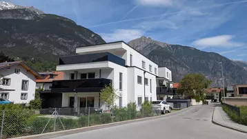 Modernes Wohngebäude mit Flachdach und Balkonen in alpiner Umgebung, umgeben von Bergen.