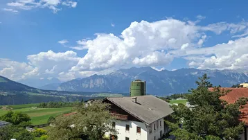 Traditionelles Haus mit Bergblick, umgeben von grünen Feldern und Bäumen unter blauem Himmel.