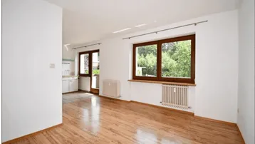 Helles Wohnzimmer mit Holzboden, weißen Wänden und Blick ins Grüne, angrenzend an eine Küche.