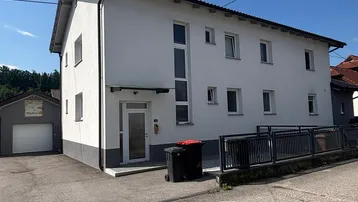 Weißes Mehrfamilienhaus mit dunklem Dach und blauer Himmel, umgeben von einer asphaltierten Zufahrt.