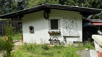 Kleines weißes Gebäude mit dunklem Dach, umgeben von einem verwilderten Garten und hohen Bäumen.