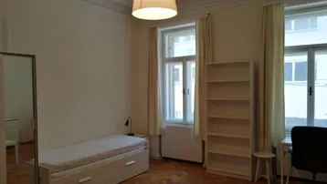 Helles Schlafzimmer mit Parkettboden, hohen Decken und großen Fenstern, ausgestattet mit einem Bett und Regal.