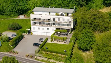 Luftaufnahme eines weißen Gebäudes mit mehreren Balkonen, umgeben von viel Grün und einem Parkplatz.
